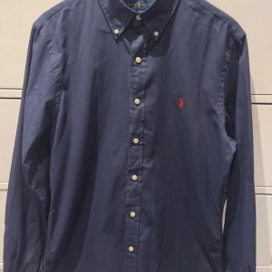 Mörkblå skjorta från Ralph Lauren - Klassisk mörkblå skjorta från Ralph Lauren med knappar framtill och ikonisk röd logga broderad på bröstet. Skjortan har lång ärm och button down-krage, perfekt för en clean och stilren look.