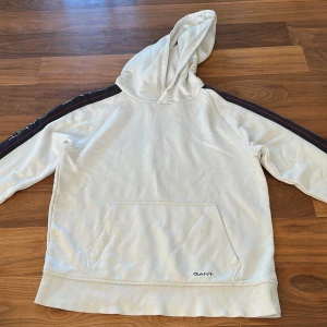 Vit hoodie från GANT med logga - Vit hoodie från GANT med marinblå och röda detaljer längs ärmarna där GANT-loggan är broderad. Klassisk känguruficka framtill och huva. Mjuk bomullskvalitet och ribbade muddar vid ärmslut och nederkant. Perfekt för en chill och sportig look.