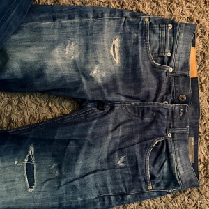 Dondup skinny fit slitna jeans - Snygga blå jeans från Ritchie i skinny fit-modell med slitningar och hål på benen för en trendig look. Jeansen har klassisk femficksdesign, silverfärgade nitar och tydliga slitdetaljer framtill. Perfekta för dig som gillar en streetig stil.