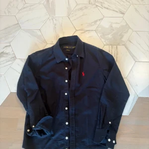 Mörkblå skjorta från Ralph Lauren - Klassisk mörkblå skjorta från Ralph Lauren med vit knappstängning och röd broderad logga på bröstet. Skjortan har lång ärm, krage och är tillverkad i mjuk bomull. Perfekt för dig som gillar stilrena och tidlösa plagg.