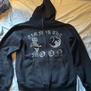 Svart hoodie Aim For The Moon L - Svart hoodie med dragkedja från Aim For The Moon. Framsidan har ett stort tryck med texten 'Aim For The Moon', en tecknad figur och en måne i silverfärgade detaljer. Utrustad med huva och två fickor framtill. 