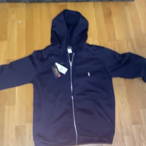 Marinblå hoodie från Polo Ralph Lauren - Snygg marinblå hoodie från Polo Ralph Lauren med dragkedja framtill och klassisk vit logga på bröstet. Tröjan har huva, långa ärmar och känguruficka. Perfekt för en chill och stilren look.