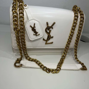 Vit YSL handväska med guldkedja - Snygg vit handväska från YSL med klassisk logga i guld framtill och matchande guldfärgad kedjerem. Väskan är i slät skinn och har en stilren, boxig form. Perfekt för dig som vill ha en lyxig accessoar med statement-detaljer.