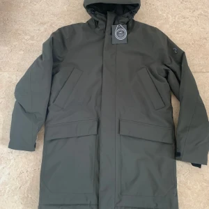Mörkgrön parkajacka med huva - Snygg mörkgrön parkajacka från Peak Performance med huva och stora fickor framtill. Jackan har en stilren design med raka linjer och är perfekt för kyliga dagar. Tillverkad i slitstarkt material och har långa ärmar samt diskret logga på ärmen.