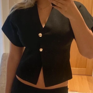Trekvartsärmad topp guldknappar från Zara - Säljer en svart topp från Zara med korta ärmar och v-ringning. Blusen har två snygga guldknappar framtill och en croppad passform. Perfekt för dig som gillar stilrena och trendiga plagg med en twist. Går inte att köpa längre ❤️
