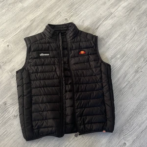 Svart dunväst från Ellesse - Snygg svart dunväst från Ellesse med quiltad design och hög krage. Västen har dragkedja framtill och broderade loggor på bröstet. Perfekt för lager-på-lager och streetstyle. Klassisk och sportig look.