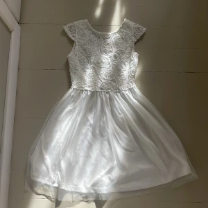 Vit spetsklänning med pärlknappar - Superfin vit kortklänning med spets och broderade detaljer upptill. Kjolen är i flera lager tunn, glansig polyester som ger ett flowy intryck. Baksidan har v-ringning och pärlknappar. Perfekt för sommarens speciella tillfällen!