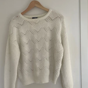 Vit stickad tröja från KappAhl - Säljer en vit stickad tröja från KappAhl med snyggt hålmönster och rund halsringning. Tröjan har lång ärm och ribbade muddar vid ärmslut och nederkant. Perfekt för lager-på-lager och en mysig stil.