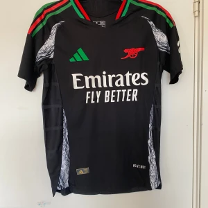 Arsenal svart bortatröja Adidas M - Svart Arsenal bortatröja från Adidas i slim fit med gröna och röda detaljer på axlarna, vitt tryck och röd kanonlogga. Tillverkad i återvunnet funktionsmaterial HEAT.RDY. Snygga mönstrade paneler längs sidorna och klassisk V-ringning.