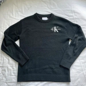 Svart stickad tröja Calvin Klein Jeans - Svart stickad tröja från Calvin Klein Jeans med broderad CK-logga på bröstet. Klassisk rund halsringning och ribbade muddar vid ärmslut och nederkant. Perfekt för dig som gillar stilrena och tidlösa plagg.