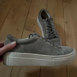 Gråa sneakers i mocka från Vagabond - Snygga grå sneakers från Vagabond i mjuk mocka med vita, platta sulor och guldfärgade öljetter. Skorna har klassisk snörning och en stilren, rund tå. Perfekta för dig som gillar en clean och enkel look med lite extra edge.