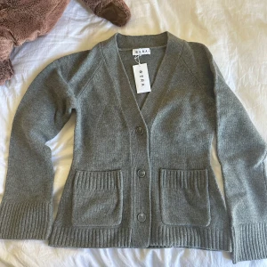 Grå kofta från Wera med fickor - Så fin och mysig grå kofta från Wera med djup v-ringning, två stora fickor framtill och breda ribbade muddar. I ull.  Aldrig använd, lappen sitter kvar. Köp för 899kr