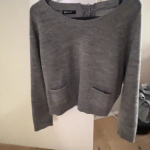 Grå stickad tröja från Gina Tricot - Mysig grå stickad tröja från Gina Tricot med två små fickor framtill och ribbade muddar. Tröjan har rund halsringning, långa ärmar och två dekorativa knappar i nacken. Perfekt för lager-på-lager under kyliga dagar.