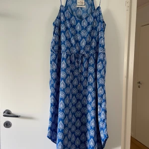 Blå mönstrad sommarklänning Holly & Whyte, Lindex - Snygg blå klänning från Holly & Whyte by Lindex med vitt mönster och smala axelband. Klänningen har knyt i midjan för en skön passform och är tillverkad i ett lätt och luftigt material, perfekt för varma dagar. Axelbandet har gått av bak på ena sidan, men bara att enkelt sy fast igen. 