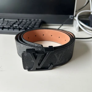 Svart Louis Vuitton skärp i skinn - Säljer ett svart Louis Vuitton bälte i skinn med klassiskt monogrammönster och stor LV-logga i matt svart metall. Insidan är ljusbrun och bältet har exklusiv känsla med tydliga detaljer och präglad märkning. Perfekt accessoar för att lyfta din outfit. Har tyvärr tappat bort kvittot. Priset är inte hugget i sten!