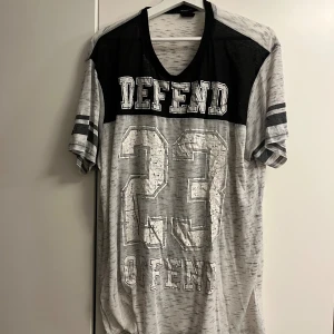 Grå och svart t-shirt FSBN XL - Sportig t-shirt från FSBN i grått och svart med tryck 'DEFEND 23 OFFEND' på framsidan. T-shirten har korta ärmar med dubbla ränder och v-ringad hals. Regular fit och tillverkad i mjukt material, perfekt för en avslappnad streetstil.