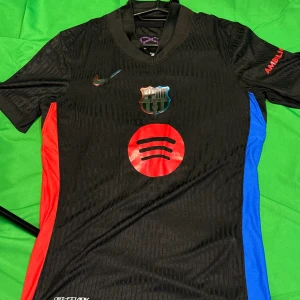 FC Barcelona Lamine Yamal svart matchtröja - Snygg svart FC Barcelona fotbollströja från Nike med Lamine Yamal och nummer 19 på ryggen. Tröjan har röda och blå sidopaneler, klubbmärke på bröstet och Spotify-logga framtill. Tillverkad i lätt och ventilerande material, perfekt för match eller träning. Liknande modell som originalet!