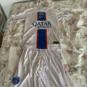 PSG Mbappé fotbollströja Nike 164 - Paris Saint-Germain fotbollströja med Mbappé och nummer 7 på ryggen. Vit med blå och röda ränder framtill, PSG-logga och Qatar Airways tryck. Kortärmad, tillverkad i Dri-FIT material som andas. Snygg design med klassiska PSG-detaljer och Nike-logga.