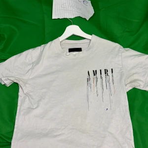 Vit AMIRI t-shirt - Snygg vit t-shirt från AMIRI med stort tryck på både bröstet och ryggen. Trycket består av AMIRI-loggan med coola färgstänk och droppar i svart, blått och orange. Klassisk passform och rund hals, perfekt för en trendig streetstyle-look.Liknande modell som originalet!