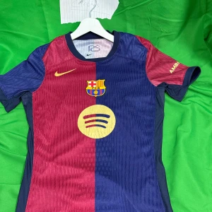 FC Barcelona Fotball T-shirt  Nike blå/röd - Säljer  FC Barcelona matchtröja från Nike med klubbmärke och Spotify-logga på bröstet. Tröjan är kortärmad och har klassiska blå och röda paneler med subtilt mönster i tyget. Materialet är lätt och andas, perfekt för fotboll eller supporterliv. Liknande modell som originalet!