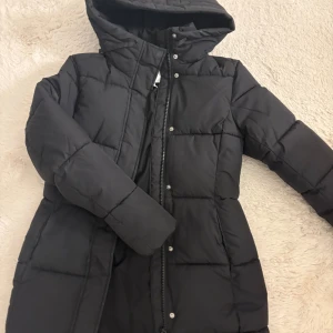 Svart lång pufferjacka från H&M - Svart lång pufferjacka från H&M med huva och dragkedja framtill. Jackan har quiltad design, tryckknappar och är perfekt för kalla dagar. Snygg och enkel stil som håller dig varm hela vintern.