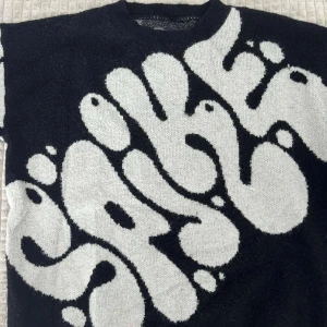 Svartvit stickad tröja med grafiskt mönster - Cool stickad tröja från Spike i svart och vitt med ett stort grafiskt mönster och texten 'SPIKE' på framsidan. Tröjan har rund halsringning och är perfekt för dig som gillar statement-plagg och streetstyle.