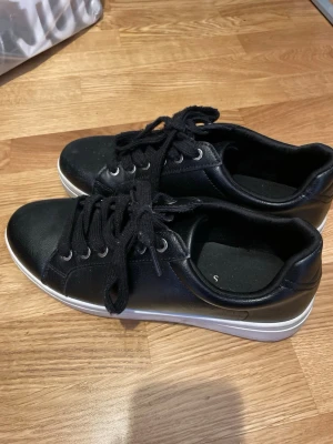 Svarta sneakers med vit sula - Bra skick, ska bara rengöras. Svarta sneakers i syntetmaterial med klassisk design och vit platt sula. Skorna har svarta snören, silverfärgade öljetter och rund tå. Perfekta för en clean och enkel look. Från Ullared i storlek 41, passar 40.