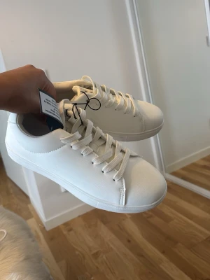Vita sneakers med snörning, storlek 37 - Fräscha vita sneakers i syntetmaterial med klassisk design och rund tå. Skorna har platt sula och vita skosnören. Perfekta för dig som gillar en clean och stilren look. Passar till det mesta och är superenkla att matcha. Skorna liknar ” Axel A*igato” och är verkligen sååå fina!!