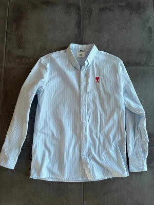 Randig skjorta från Ami i bomull - Säljer en snygg ljusblå och vit randig skjorta från Ami Paris. Skjortan har klassisk krage, långa ärmar och knäppning framtill. På bröstet finns Ami:s ikoniska röda hjärta. Perfekt för dig som gillar stilrena detaljer.