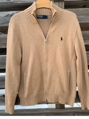 Beige stickad kofta från Polo Ralph Lauren - Säljer en beige stickad kofta från Polo Ralph Lauren med ribbad hög krage och dragkedja framtill. Klassisk marinblå logga broderad på bröstet och två fickor. Perfekt till lager på lager under höst och vinter.