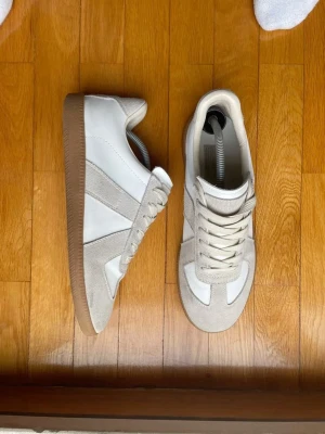 Maison margiela sneakers - Säljer ett par stilrena maison margiela gats i vitt och beige med ovandel i skinn och detaljer i mocka. Skorna har beige snörning, gummisula och en klassisk, låg siluett. Perfekta för dig som gillar minimalistisk och clean stil.