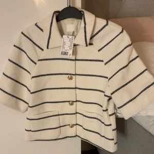 Randig overshirt från H&M - Kort, krämvit overshirt från H&M med svarta horisontella ränder och fyra guldknappar framtill. Jackan har korta ärmar, två fickor fram och en klassisk krage. Materialet är strukturerat och känns lite tjockare, perfekt för lager på lager.