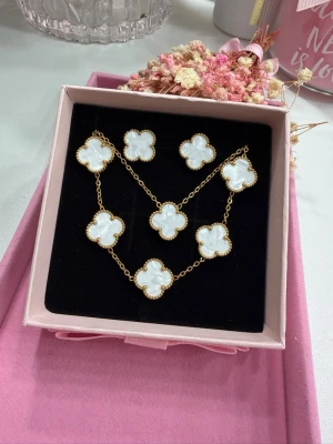 Klöver-set - Elegant och stilrent klöver-set i vacker vit färg som ger en lyxig och trendig känsla. Setet består av halsband, armband och örhängen, tillverkat i högkvalitativt rostfritt stål med exklusiv 18K guldplätering. Klöverdesignen i 15 mm ger ett tidlöst och feminint uttryck som passar både vardag och fest. Armbandet har en längd på 17,5 cm till första ringen samt 5 cm förlängningskedja för optimal och justerbar passform. Setet levereras i en elegant ask, perfekt som present.