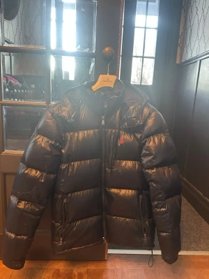 Mörkblå pufferjacka från Polo Ralph Lauren - Säljer en mörkblå  pufferjacka från Polo Ralph Lauren med huva och dragkedja. Jackan har en klassisk quiltad look, två sidofickor med dragkedja och en bröstficka. Materialet är glansigt och fylligt, perfekt för kalla dagar.