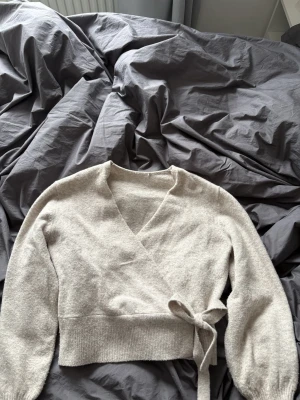 Beige omlottstickad kofta med knyt - Mysig beige omlottkofta i stickat material med bred ribbad kant och knytband i midjan. Tröjan har lång ärm och v-ringad hals, perfekt för lager på lager under höst och vinter. Enkel och stilren design som är lätt att matcha. Säljer den då den inte används längre❣️