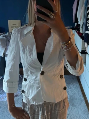 Vit kavaj med stora knappar - Säljer en vit kavaj med markerad midja och stora mörka knappar framtill och på ärmsluten. Jackan har klassisk krage och trekvartsärmar. Perfekt för dig som vill ha en stilren och fräsch look.