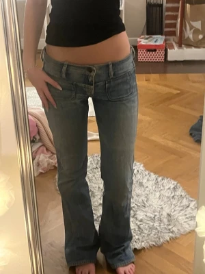 Lågmidjade bootcut jeans blå - Säljer ett par riktigt snygga lågmidjade jeans i ljusblå tvätt med bootcut-ben. Jeansen har klassiska fem fickor, bälteshällor och en cool vintagekänsla. Perfekta för dig som gillar Y2K-stil och vill ha ett par jeans som sitter lågt på höften. Midjemåttet är 39cm och innerbenslängden är 82! Jag är 170 som referens. Lite slitna längst ner (se bild 5)