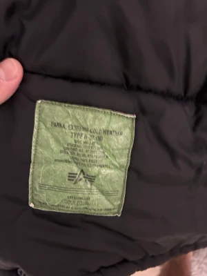 Svart Alpha Industries parkajacka med fuskpäls - Parkajacka från Alpha Industries i svart med snygg brun fuskpäls på huvan. Jackan har dragkedja, flera fickor och detaljer som röd tag på ärmen. Materialet är nylon och polyester, perfekt för kalla vinterdagar.