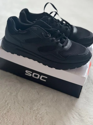 Svarta sneakers från SOC, storlek 46 - Säljer ett par helsvarta sneakers från SOC i storlek 46. Skorna har en sportig look med meshpaneler och detaljer i syntetmaterial. Klassisk snörning och rejäl yttersula med greppvänligt mönster. Perfekta till vardags eller träning.