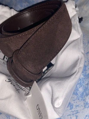 Brunt mockabälte från ONEWear - Säljer ett stilrent brunt bälte från ONEWear i mjuk mocka eftersom att den är förlitet. Bältet har en klassisk design med enkel metallspänne och kommer i en snygg tygpåse. Perfekt accessoar för att lyfta din outfit. 