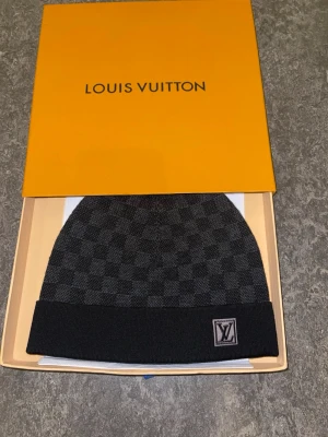 Rutig svart mössa från  - Snygg svart mössa från med diskret rutigt mönster framtill. Tillverkad i mjuk ullblandning med elastan för extra komfort. Perfekt accessoar för dig som vill ha en exklusiv och stilren look.