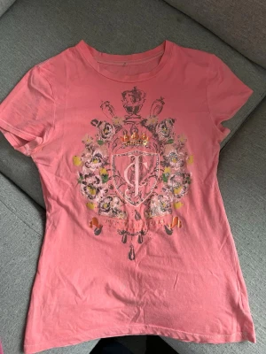 Rosa Juicy Couture t-shirt med tryck - Snygg rosa vintage t-shirt från Juicy Couture med stort tryck framtill i form av blommor och emblem. T-shirten har rund hals, korta ärmar och är gjord i mjuk bomull. Perfekt för dig som gillar statement-plagg och vill sticka ut lite extra.