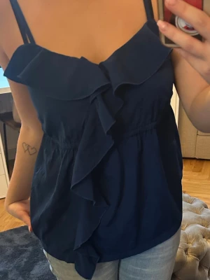 Blå volangtop från Vero Moda - Säljer en marinblå linnetopp från Vero Moda med breda axelband och snygg volangdetalj framtill. Toppen har en luftig passform och är gjord i mjuk polyester, perfekt för varma dagar. Enkel att matcha med jeans eller kjol för en chill look.