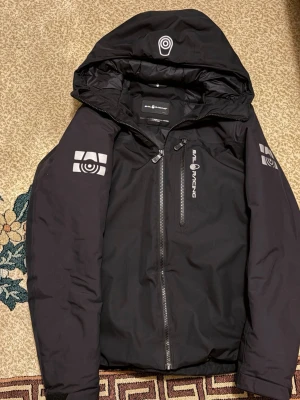 Svart Sail Racing jacka med huva - Svart jacka från Sail Racing med huva och coola loggor på ärmar och bröst. Jackan har dragkedja framtill och på bröstfickan, samt sportig design. Perfekt för dig som gillar stilrena och funktionella ytterplagg.☀️