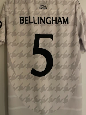Real Madrid Bellingham matchtröja - Säljer en Real Madrid matchtröja med Bellingham och nummer 5 på ryggen. Tröjan är vit med ljusgrått rutigt mönster, broderat klubbmärke och svarta Adidas-detaljer. Materialet är polyester och modellen är kortärmad med rund hals.