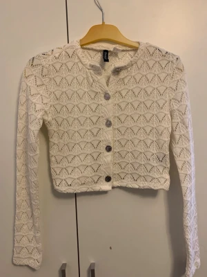 Vit stickad kofta från Divided - Säljer en söt vit stickad kofta från Divided med genombrutet mönster och snygga silverfärgade knappar. Koftan är croppad och har långa ärmar, perfekt att slänga över en topp för en chill look.