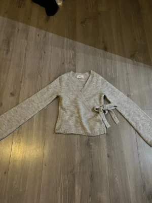 Grå omlottkofta med knyt - Mysig grå omlottkofta med knyt i sidan och långa ärmar. Tröjan har en ribbad kant nedtill och vid ärmsluten, vilket ger en snygg och enkel look. 