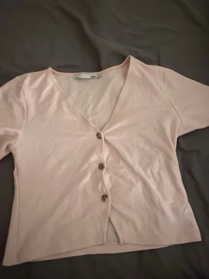 Ljusrosa blus med v-ringning - Säljer en ljusrosa blus från 157 med v-ringad hals och tre dekorativa knappar framtill. Blusen har korta ärmar och är tillverkad i mjuk bomull, perfekt för vår och sommar. Enkel och stilren design som är lätt att matcha med olika outfits.