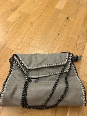Grå axelväska med kedjedetaljer - Snygg Stella McCartney inspirerad axelväska i mockaimitation med coola kedjor som axelrem och dekorativa sömmar längs kanterna. Väskan har lockstängning och flera innerfack med dragkedja för smart förvaring. Perfekt för dig som gillar en stilren men edgy look.