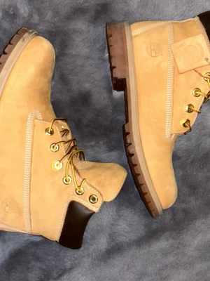 Timberland boots i beige mocka - Klassiska Timberland boots i beige mocka med robust brun sula och guldfärgade metalldetaljer. Skorna har snörning med gul- och brunmönstrade skosnören samt mörkbrunt parti vid ankeln. Perfekta för som vill ha varma och sköna skor. Skorna är storlek 40 och dom är typ helt nya dom är bara andvända några få gånger 